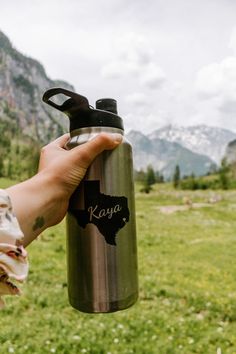 a person holding a stainless steel water bottle in front of a grassy field with mountains