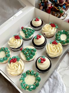 twelve decorated cupcakes in a white box on a table next to christmas decorations