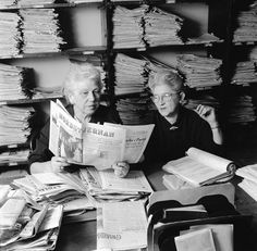 two women sitting at a table with papers in front of them and one reading the paper