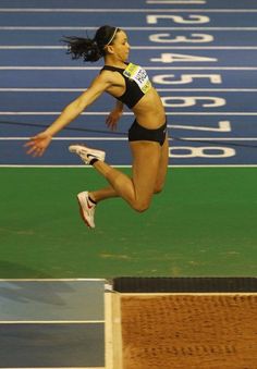 a woman jumping in the air on top of a blue and green track with an olympic sign behind her