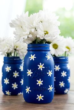 three blue mason jars with white daisies in them sitting on a wooden table next to a window