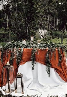 an orange and white table cloth with greenery on it is set up in the woods