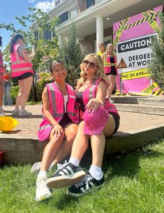 two women in pink dresses sitting on steps with construction signs behind them and people standing around