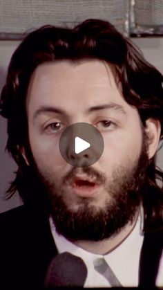 a man with long hair and beard wearing a black suit is talking into a microphone