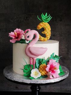 a cake decorated with pink flamingos and pineapples on a marble stand in front of a black wall