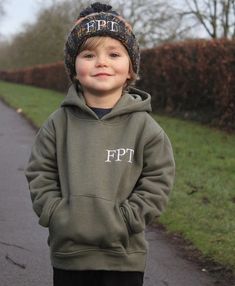 a young boy standing on the side of a road wearing a green sweatshirt and hat