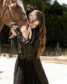 a woman in a black dress standing next to a brown horse with a bridle on it's head