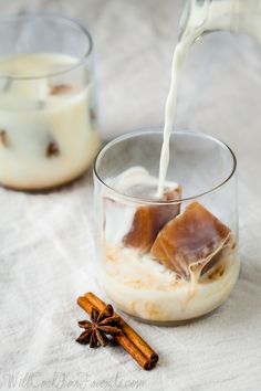 two glasses filled with pudding and milk being poured into one glass, cinnamon sticks on the other