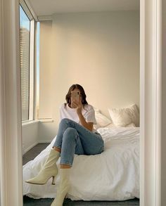 a woman sitting on top of a white bed in a bedroom next to a window