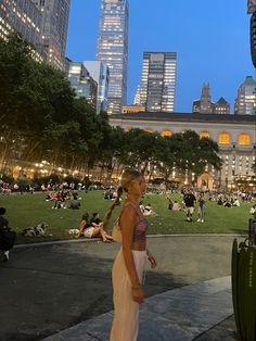 a woman standing in the middle of a park with lots of people sitting and walking around