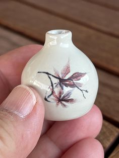 a hand holding a small white vase with leaves painted on it's side, in front of a wooden surface