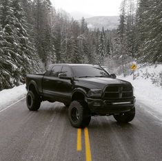 a large black truck driving down a snow covered road next to pine trees and evergreens