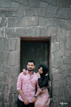 two people standing next to each other in front of a stone wall with a window