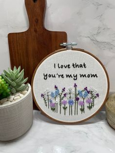 a small potted plant sitting next to a cross stitch hoop with the words i love that you're my mom on it