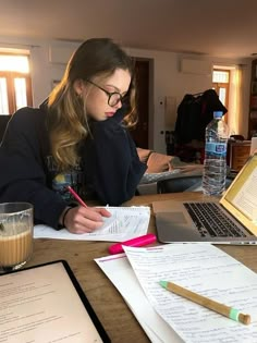 a woman sitting at a table with papers and a laptop on it, writing in front of her