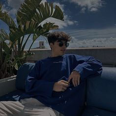 a young man sitting on top of a blue couch next to a plant and wearing sunglasses