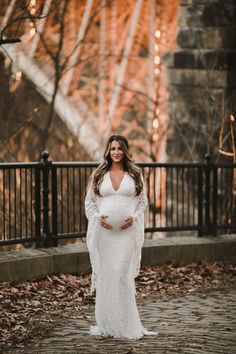 a pregnant woman standing in front of a bridge wearing a white gown and holding her belly