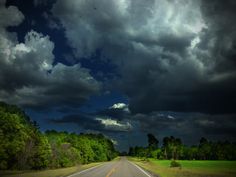 an empty road surrounded by green trees under a dark sky with storm clouds above it