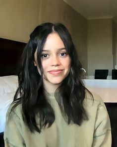 a woman with long black hair sitting in front of a bed and looking at the camera