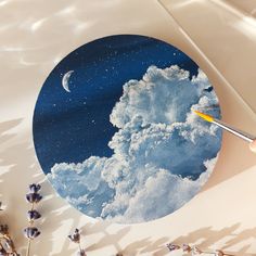 a person is holding a paintbrush and painting the clouds on a round piece of paper