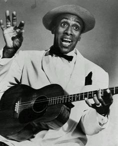 a black and white photo of a man holding a guitar in his right hand while wearing a hat