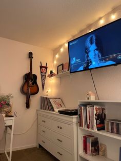 a room with a guitar, bookshelf and television on the wall in it