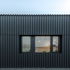 two windows on the side of a black metal building with gravel ground and sky in background