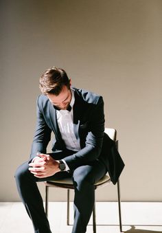 a man in a tuxedo sits on a chair and looks down at his watch