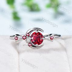 a ring with a red stone in the middle and white diamonds around it, on top of a cushioned surface