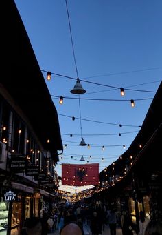 people are walking down an alley way with lights strung from the ceiling and buildings in the background