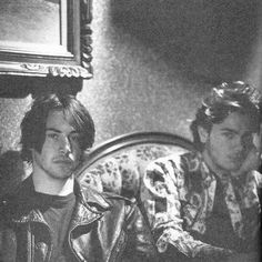 black and white photograph of two men sitting on a couch in a room with wallpaper