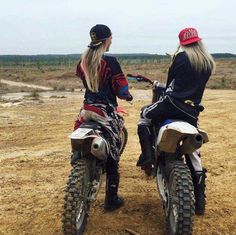 two women are sitting on dirt bikes in the middle of an open field, one is wearing a red hat