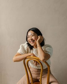 a woman sitting on top of a wooden chair next to a white wall and smiling