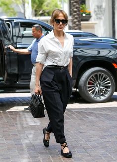 a woman in white shirt and black pants walking down the street with her handbag