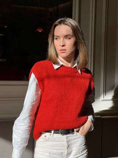 a woman standing in front of a window wearing a red sweater and white shirt with sleeves down