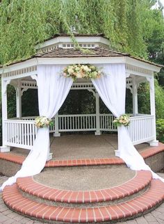 a gazebo with white drapes and flowers on the top is surrounded by brick steps