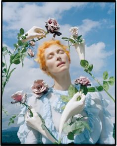 a woman with red hair and white gloves holding flowers in front of her face while looking up at the sky