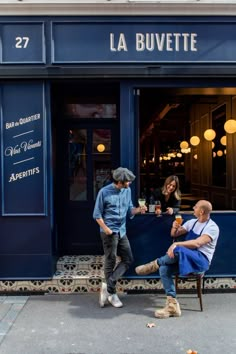 two people sitting at a table in front of a blue store with lights hanging from the windows