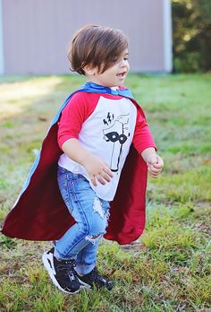 a little boy dressed in a red and white shirt with a blue cape is running through the grass