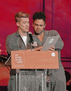 two men standing next to each other while looking at an electronic device in front of a microphone