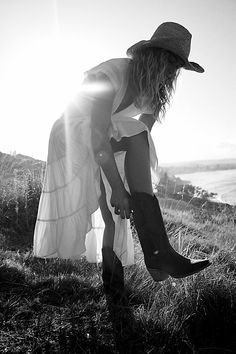 a woman in a dress and cowboy hat bending down to pick up her boots from the grass