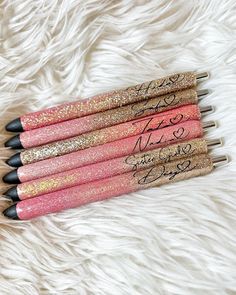 six pink and gold glitter pens with writing on them sitting on a white furry surface