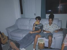 two young men sitting on a couch in a living room with guitars and other furniture