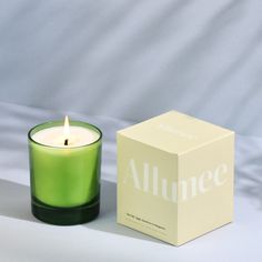 a green candle sitting next to a box on a white tablecloth with the word alumee printed on it