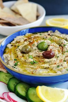 a blue bowl filled with hummus and olives next to sliced cucumbers