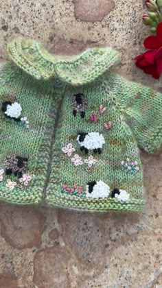 a knitted sweater with sheeps on it next to a red flower and stone wall