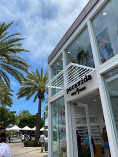 people are walking in front of a store with palm trees and blue sky behind it