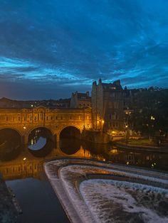 an old bridge over a river at night with buildings in the background and water flowing under it