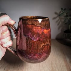 a hand holding a coffee mug on top of a wooden table next to a potted plant