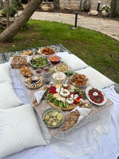 a table full of food sitting on top of a white blanket next to a tree